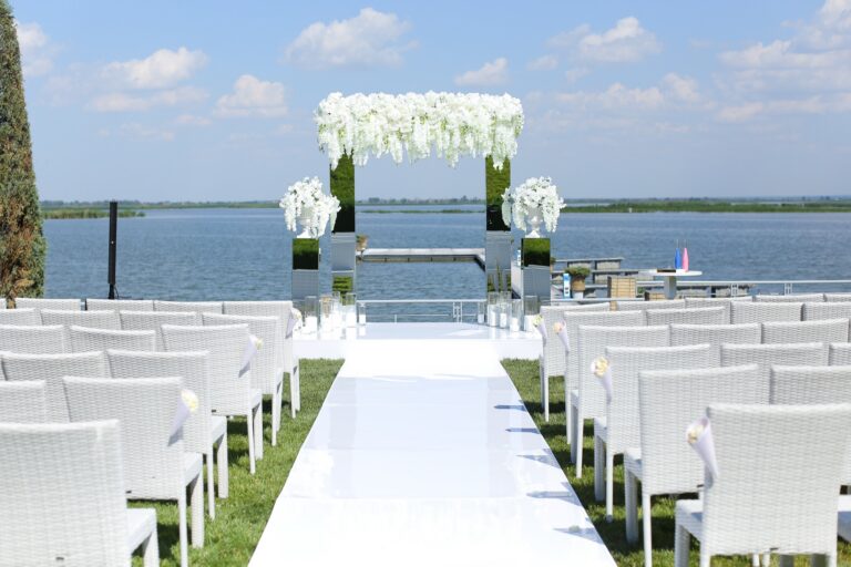 A floral wedding arch and white chairs set up for a ceremony beside a lake